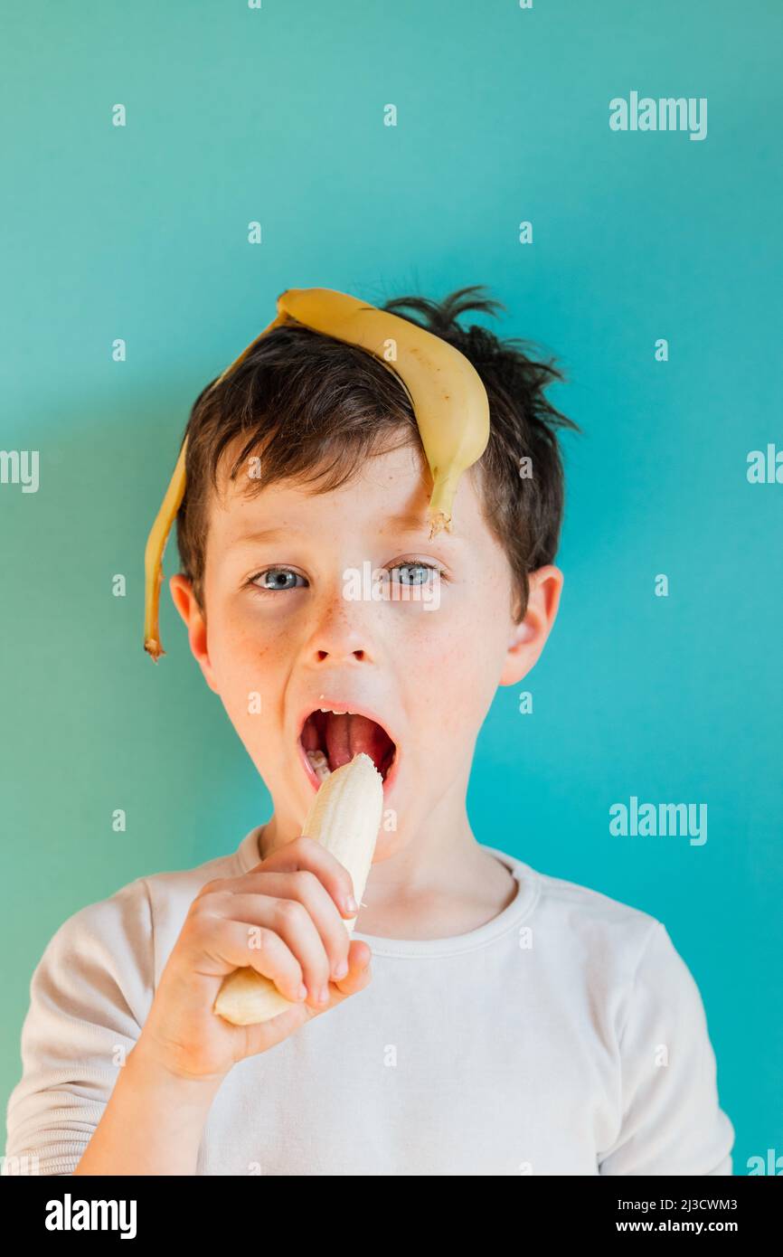 Headshot von fröhlichem Jungen mit Bananenschale auf dem Kopf und Essen von Banane, während Sie mit einem Lächeln auf blauem Hintergrund in einem hellen Studio auf die Kamera blicken Stockfoto