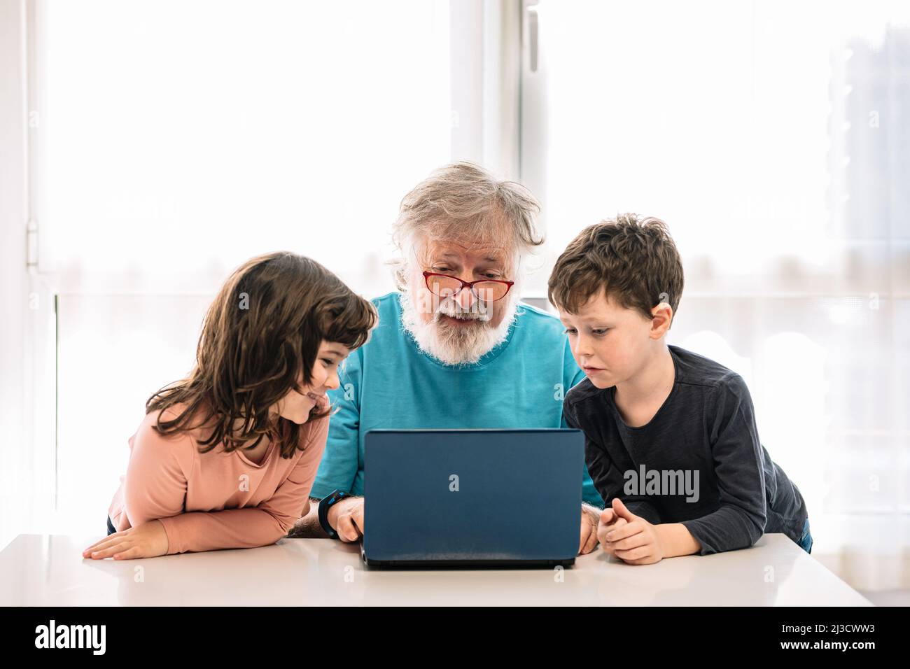 Bärtiger Großvater in einer Brille mit positiven Kindern, die sich ein Interessantes Video auf Netbook ansehen, während sie zu Hause am Tisch im hellen Raum sitzen Stockfoto