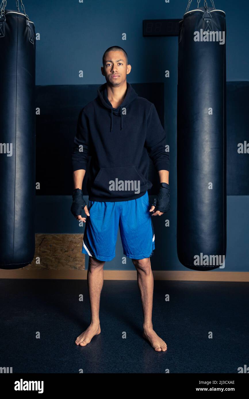 Barfuß-voller junger hispanischer Sportler in Kapuzenpullover und Shorts, der die Kamera anschaut, während er zwischen Boxsäcken im Boxstudio steht Stockfoto