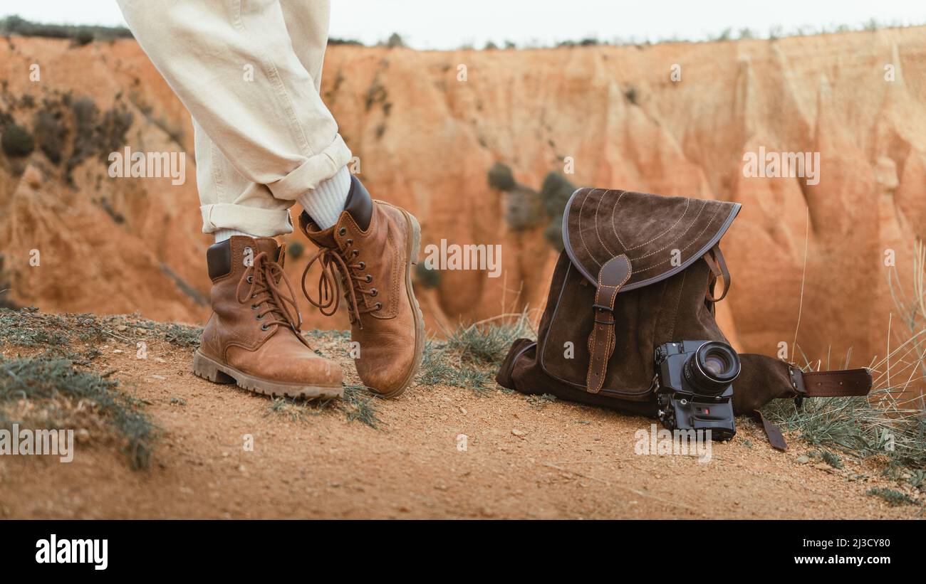 Nicht erkennbarer Mann in weißen Hosen und Lederschuhen, der in der Nähe von Wildlederrucksack und Fotokamera gegen den unartigen Canyon in der Natur steht Stockfoto