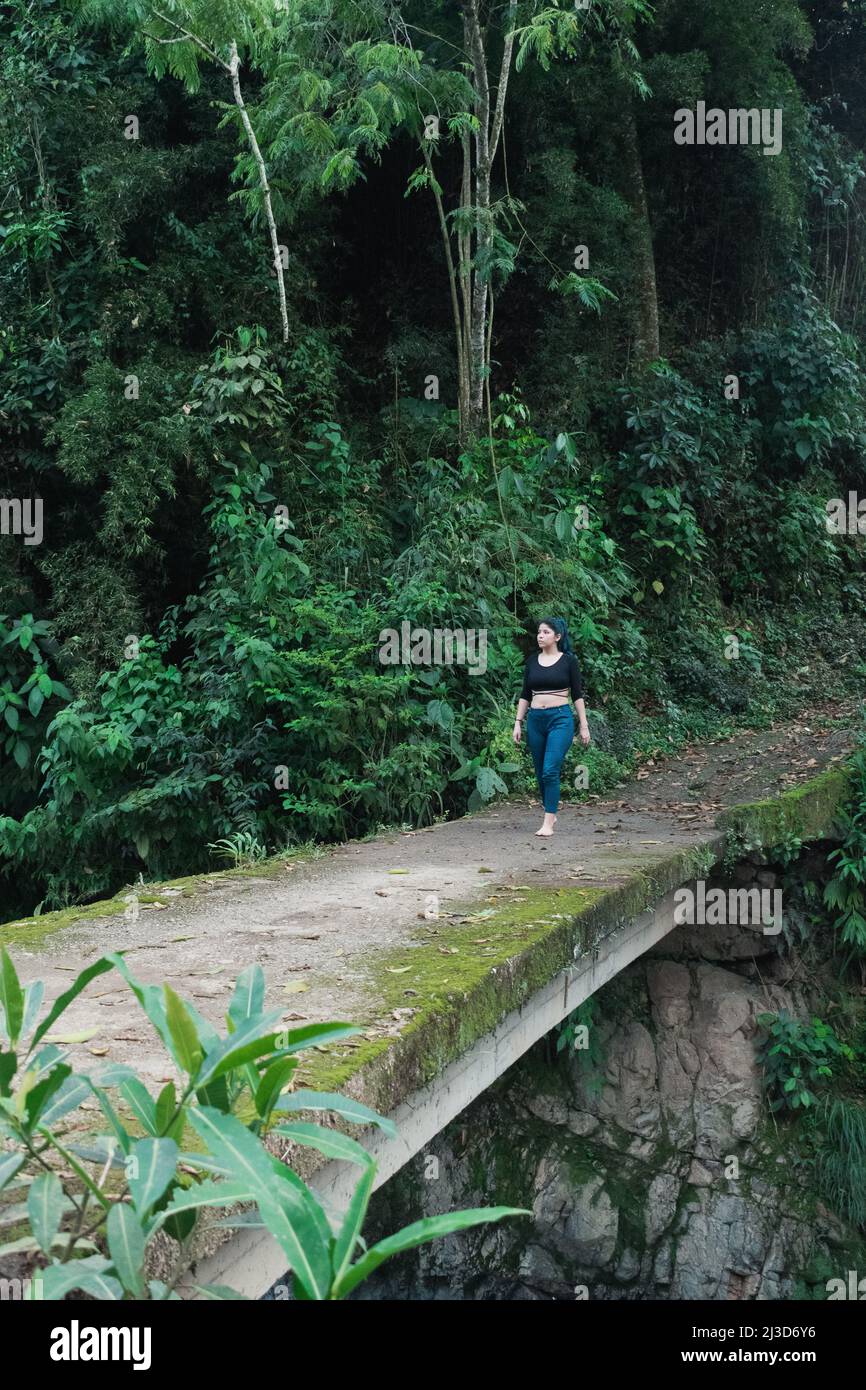 Schöne lateinerin, die mitten im kolumbianischen Dschungel eine mit grünem Moos bedeckte Zementbrücke überquert und die Natur frei und unverblümend beobachtet Stockfoto