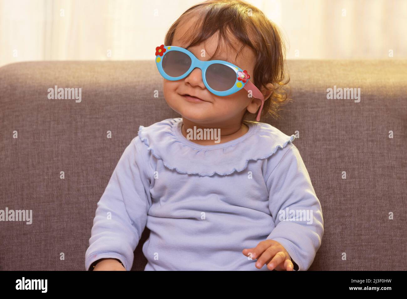 Porträt eines niedlichen Babys, das Sonnenglas trägt, während es im Wohnzimmer auf dem Sofa sitzt Stockfoto