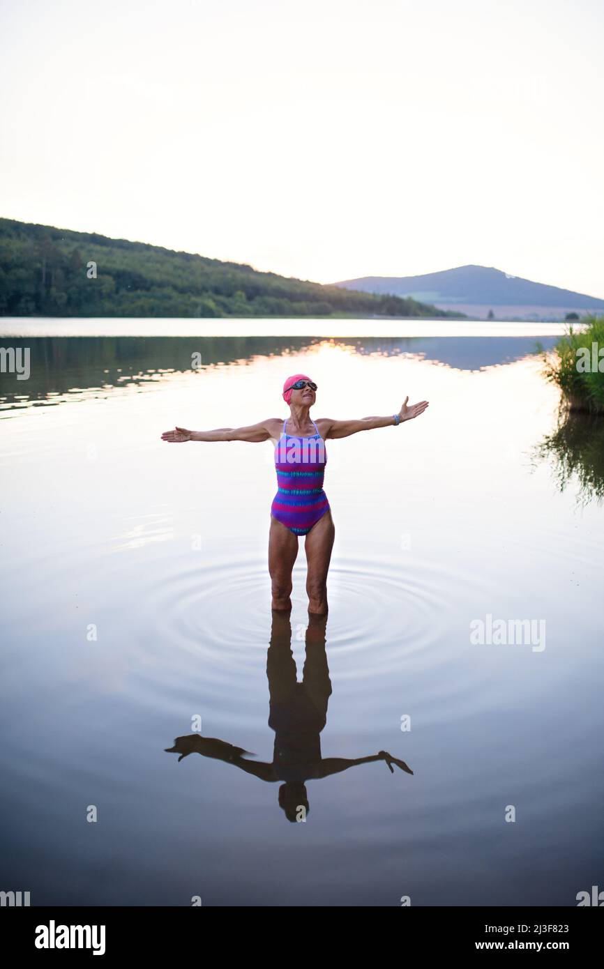 Porträt einer aktiven älteren Schwimmerin, die im Freien im See steht und sich ausdehnt. Stockfoto