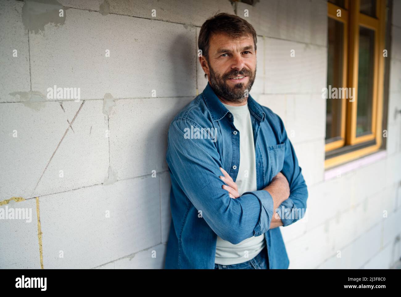 Glücklicher reifer Mann, der vor seinem neuen Haus im Bau steht und die Kamera anschaut. Stockfoto