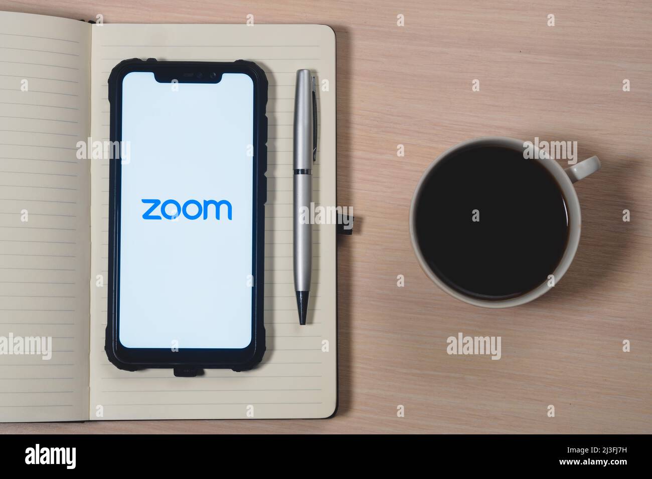 Zoom-App-Logo auf dem Bildschirm des Smartphones. Zoom Video Communications ist ein Unternehmen, das Remote-Konferenzdienste anbietet. Tagesplaner, Stift und Becher Stockfoto