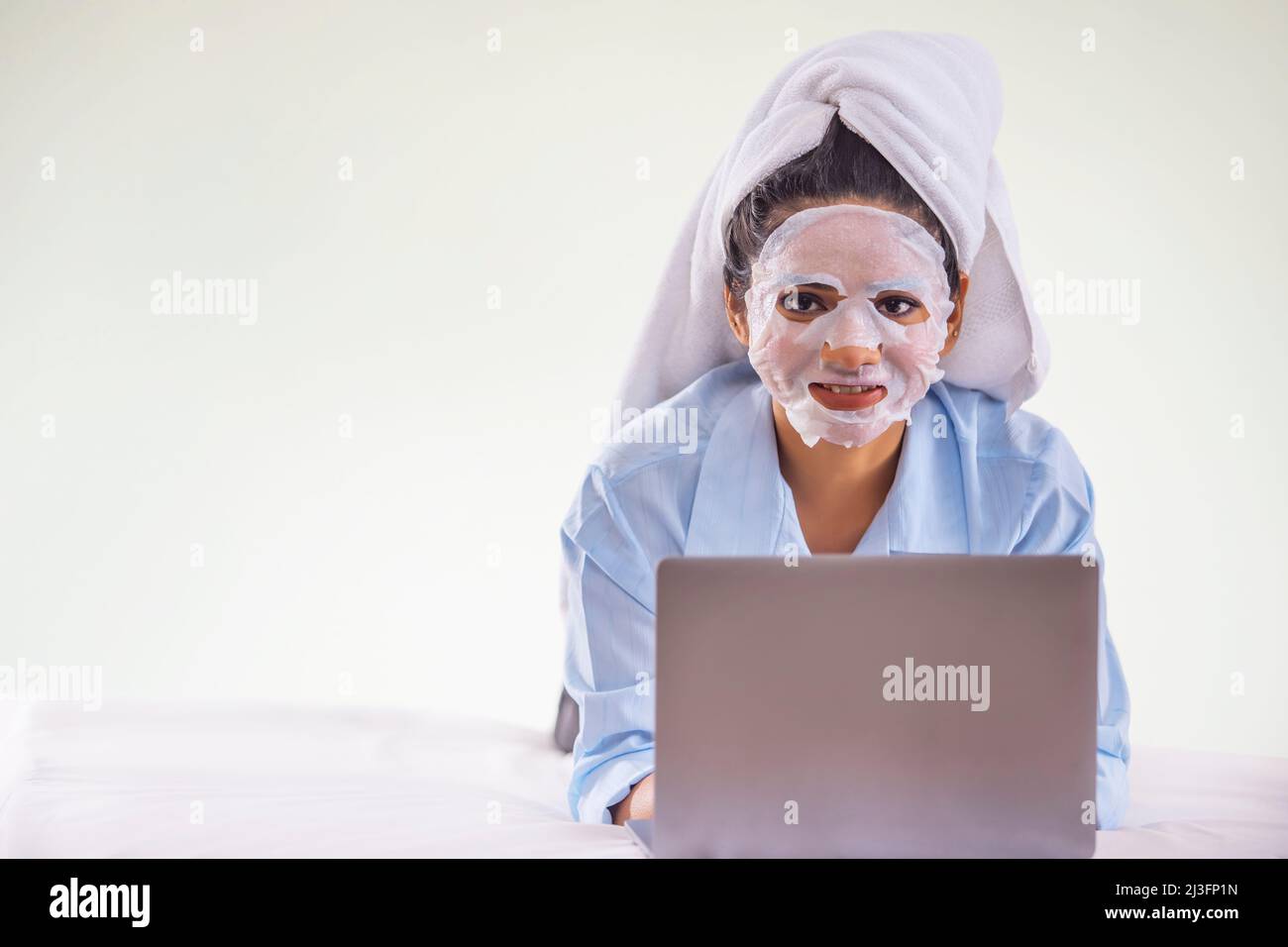 Porträt einer jungen Frau, die während der Nutzung eines Laptops im Schlafzimmer eine Gesichtsmaske auf ihr Gesicht aufträgt Stockfoto