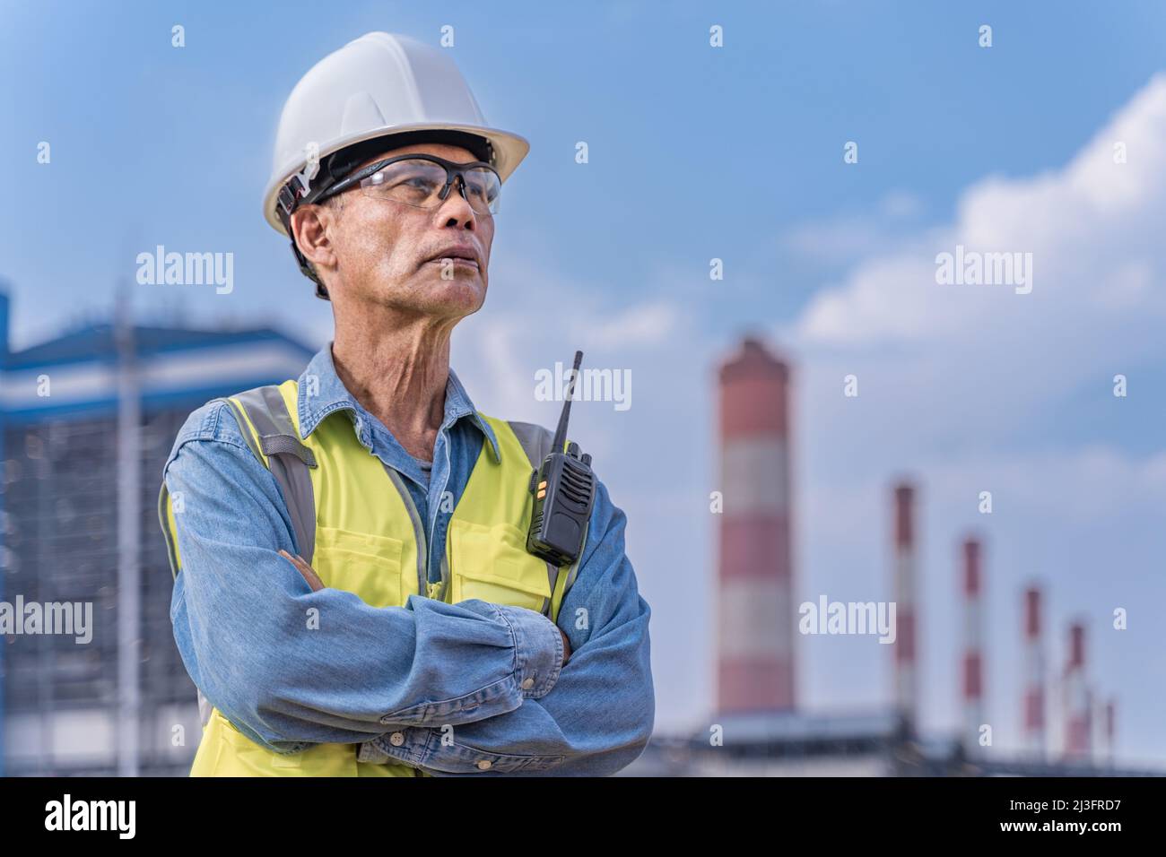 Leitender Ingenieur in seinen Arbeitstüchern mit weißem Hut, Schutzbrille und Walkie-Talkie-Radio, der am Standort des Kraftwerks stand, um sein Projekt zu überwachen Stockfoto