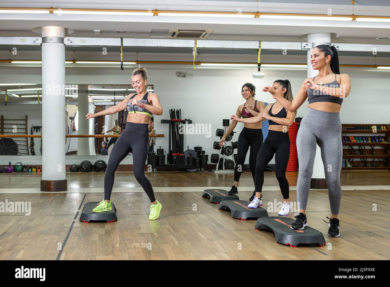 Eine Gruppe von glänzenden Sportlerinnen und Trainerin, die beim Aerobic-Training in einem geräumigen, modernen Fitnessstudio lächelnd und energisch auf Steppern tanzt Stockfoto