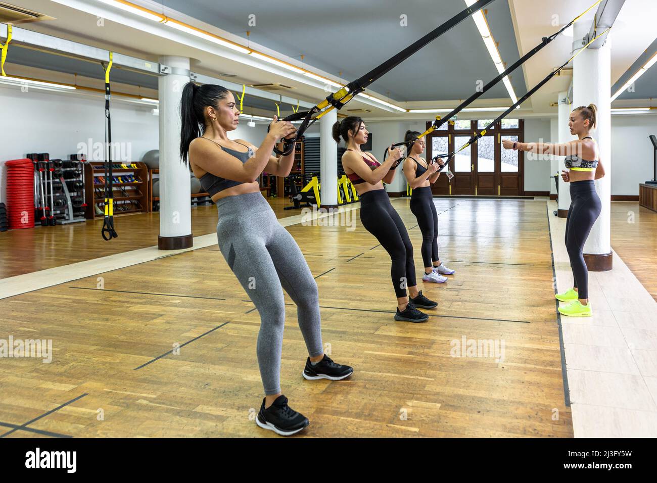 Die Trainerin erklärt Sportlerinnen, die während des Aufhängetrainings in einem geräumigen, leichten Fitnessstudio an TRX-Seilen hängen, die Übungen Stockfoto
