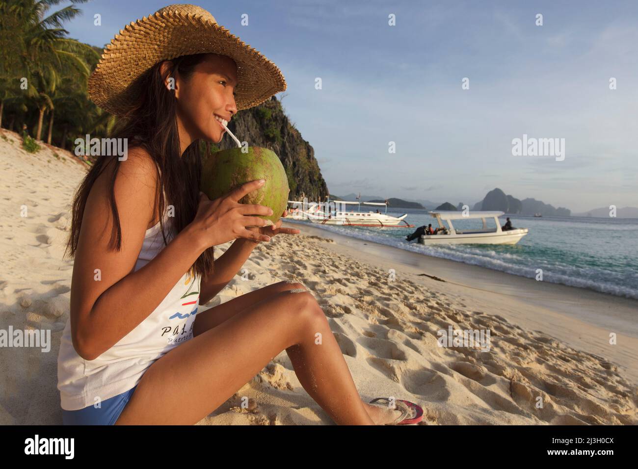 Philippinen, Palawan, El Nido, Bacuit-Archipel, Seven Commandos Beach, Eine junge philippinische Frau sitzt am Strand und trinkt aus einer Kokosnuss mit einem Stroh Stockfoto