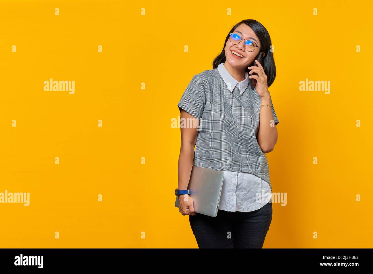 Schöne asiatische Frau hält Laptop und spricht auf Handy über gelben Hintergrund Stockfoto