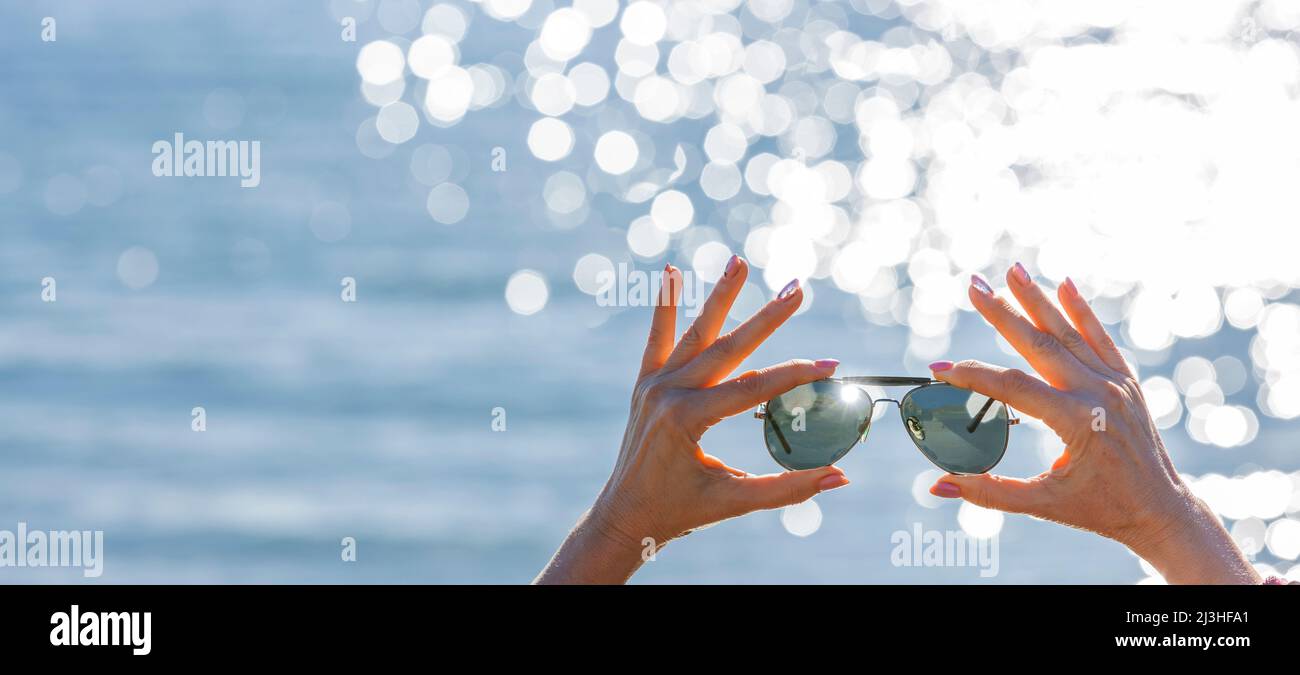 Frau Hände halten Sonnenbrille vor verschwommenem Hintergrund mit Bokeh Reflexionen Stockfoto