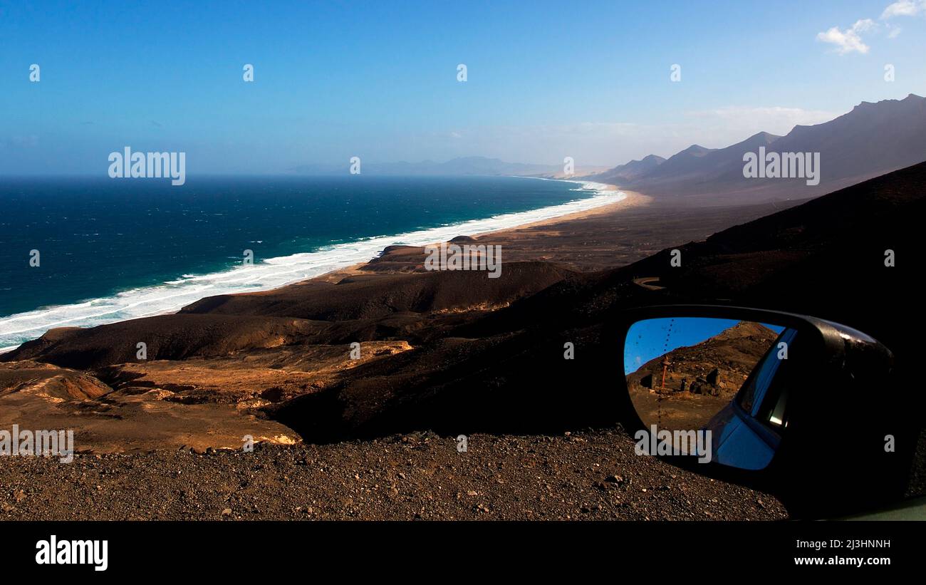 Spanien, Kanarische Inseln, Fuerteventura, südwestliche Spitze, karge Landschaft, Blick von oben auf den einsamen Nordteil der Südspitze, rechter Wagenspiegel im Bild, Gebirgskette im Hintergrund, Meer mit starker Brandung, einsamer Strand Stockfoto