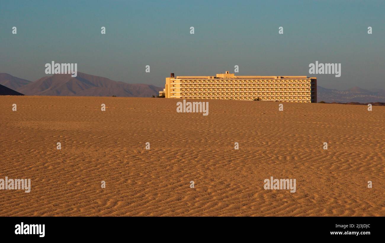 Spanien, Kanarische Inseln, Fuerteventura, Dünengebiet El Jable, Abendlicht, Sand und Düne, großes sandfarbenes Hotelgebäude, karge Hügel, blauer Himmel mit Schleierwolken Stockfoto