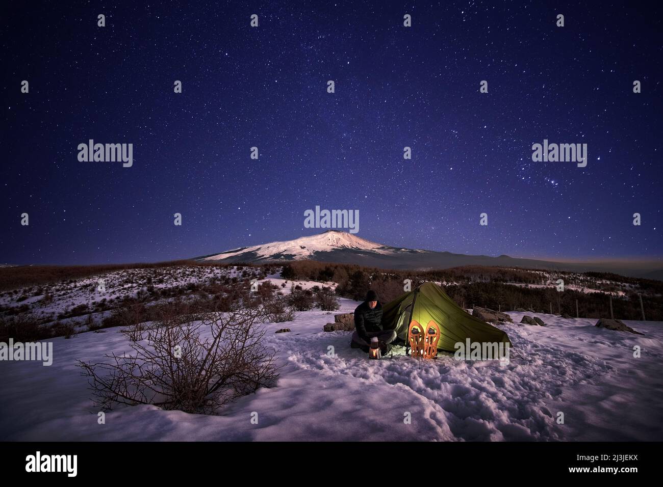 Mann, der sich in der Nähe seines beleuchteten Zeltes nachts unter Sternenhimmel auf dem Schnee auf dem Hintergrund des Ätna-Berges aus dem Nebrodi-Park, Sizilien, ausruhte Stockfoto
