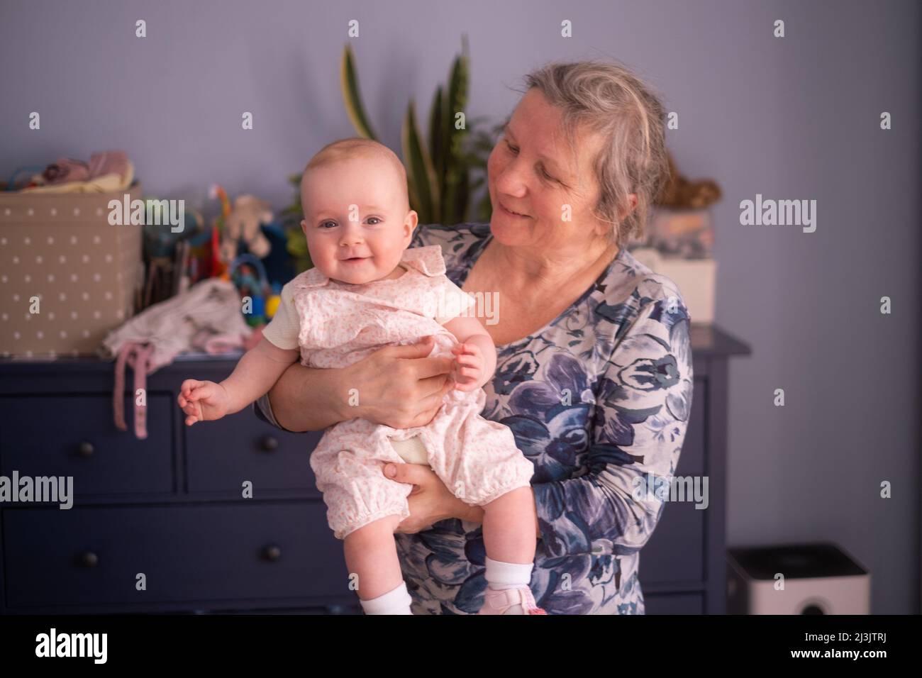 Ältere Großmutter, die zu Hause ein 7 Monate altes Baby in den Armen hält Stockfoto