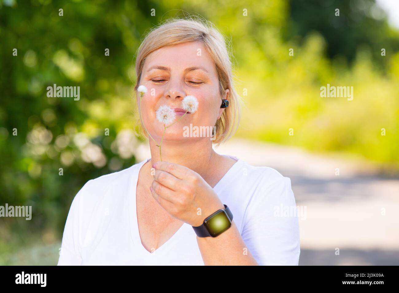 Nahaufnahme einer glücklichen blonden Frau im Freien, die eine Digitaluhr und ein Kopftelefon trägt, die in der Hand die Delandelionsblume hält und lächelt. Stockfoto
