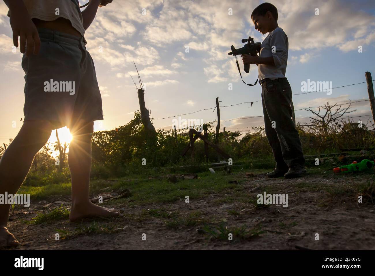 Wenn die Sonne hinter einem Stacheldrahtzaun untergeht, hält ein kleiner Junge eine Spielzeugpistole in der Hand: Cienfuegos, Kuba Stockfoto