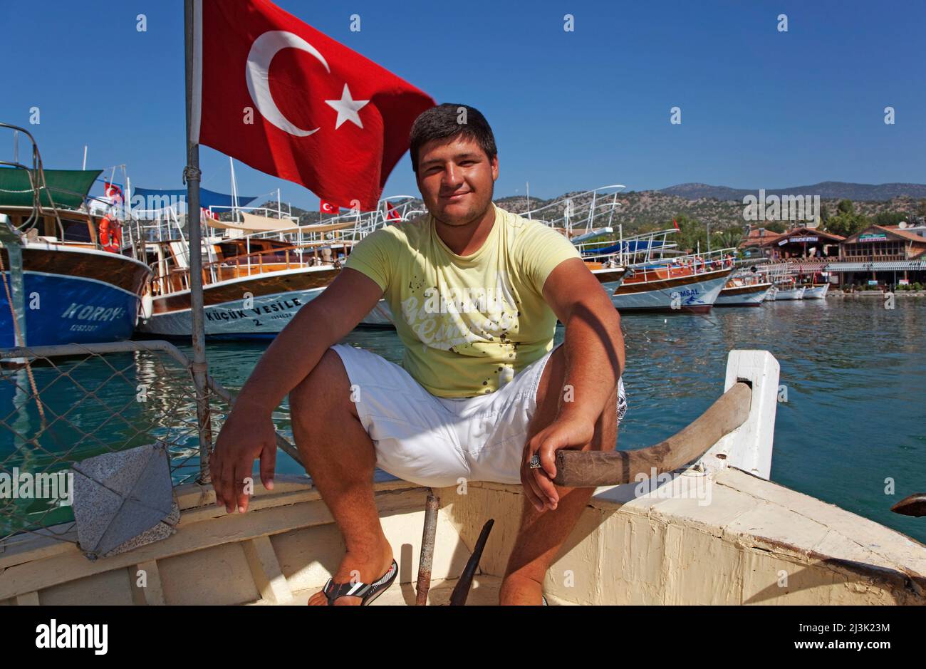 Bootsmann in Ucagiz, nahe Kas, Türkei, ein junger Mann, der am Bug eines Bootes neben einer türkischen Flagge sitzt; Ucagiz, Türkei Stockfoto