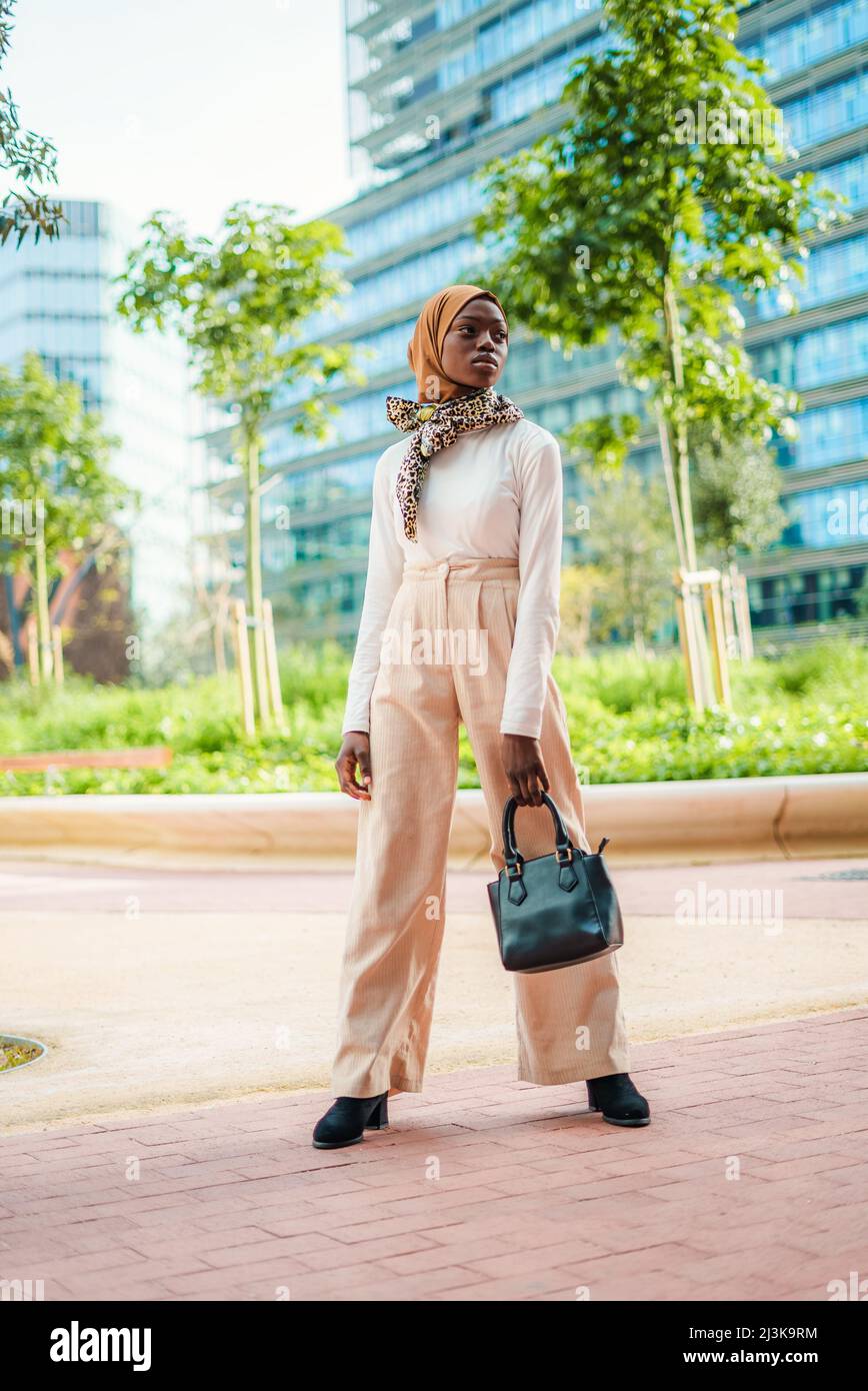 Voller Körper von selbstbewussten Frauen, gekleidet in schickes Freizeitoutfit und traditionellen islamischen Hijab mit Schal am Hals, der im Park steht und wegschaut Stockfoto