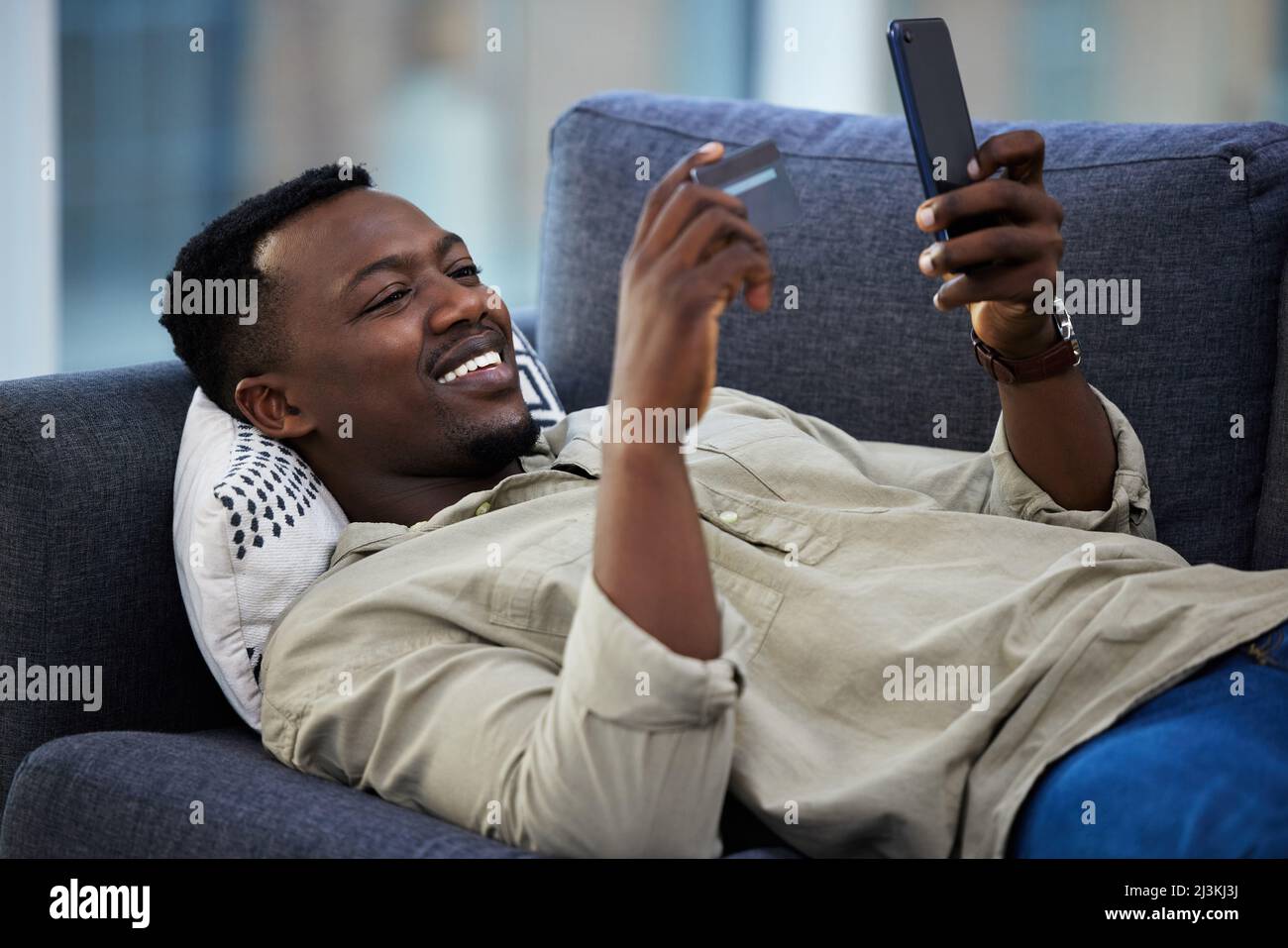 Einkaufen von der bequemen Couch aus. Aufnahme eines jungen Mannes mit Kreditkarte und Telefon auf der Couch zu Hause. Stockfoto
