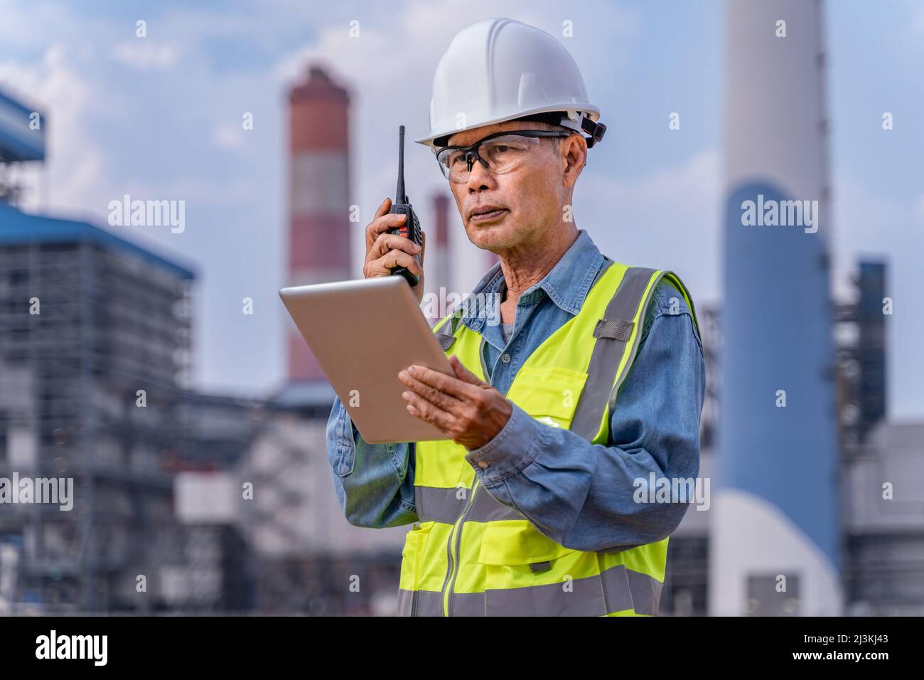 Leitender Supervisor-Ingenieur in seinen Arbeitstüchern mit weißem Hut, Schutzbrille, die sein Walkie Talkie-Radio benutzte, um während des Urlaubs mit seinem Team zu kommunizieren Stockfoto