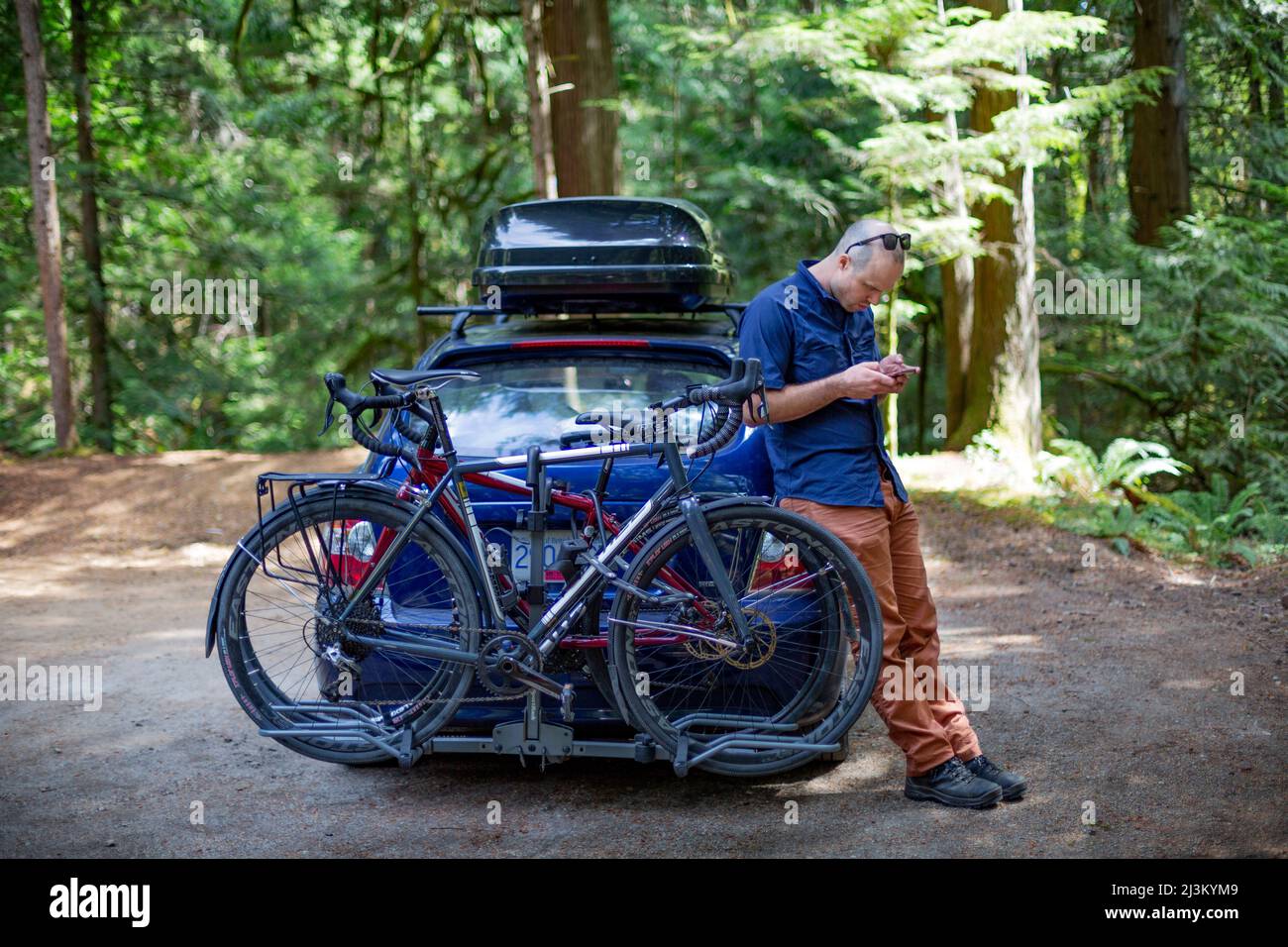 Der Mann steht mit seinem Smartphone gegen sein Auto gelehnt, mit dem Fahrrad auf dem Fahrradträger und dem Dachträger auf dem Auto; British Columbia, Kanada Stockfoto