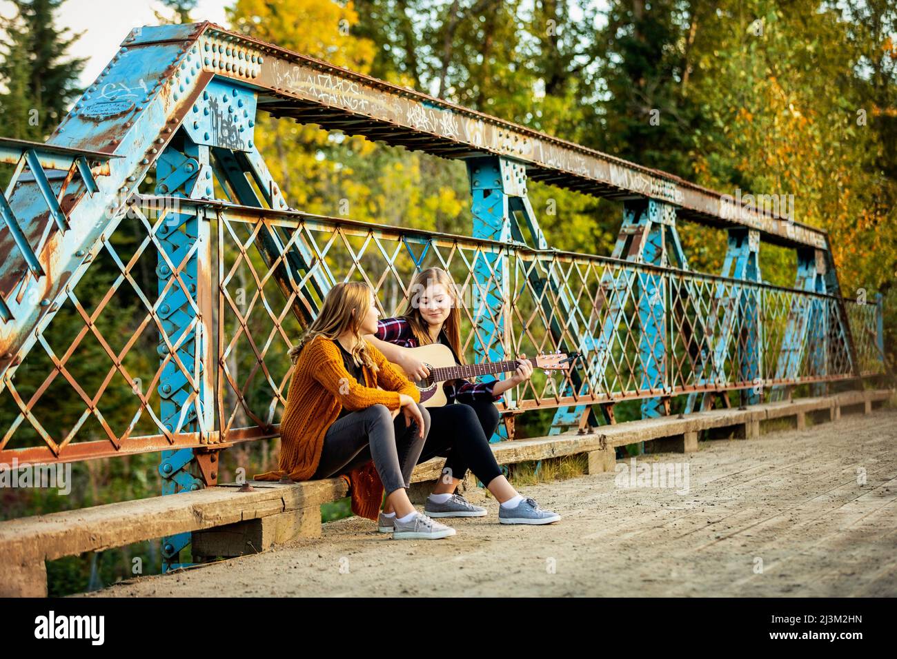 Zwei Schwestern sitzen auf einer Parkbrücke, eine mit akustischer Gitarre, und genießen gemeinsam Musik; Edmonton, Alberta, Kanada Stockfoto