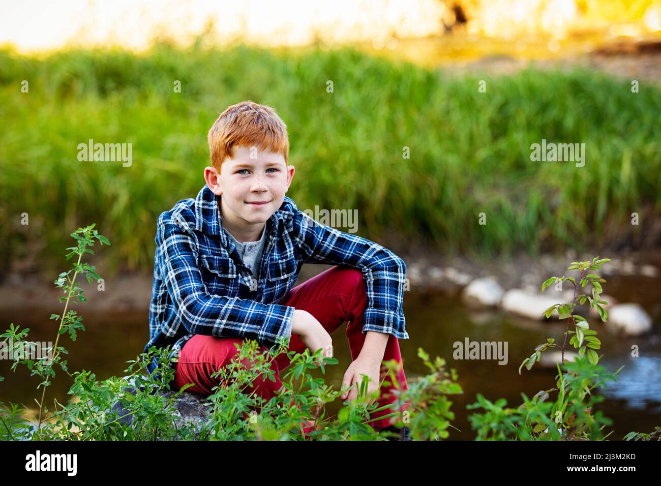 Außenportrait eines Jungen mit roten Haaren, der neben einem Bach sitzt; Edmonton, Alberta, Kanada Stockfoto
