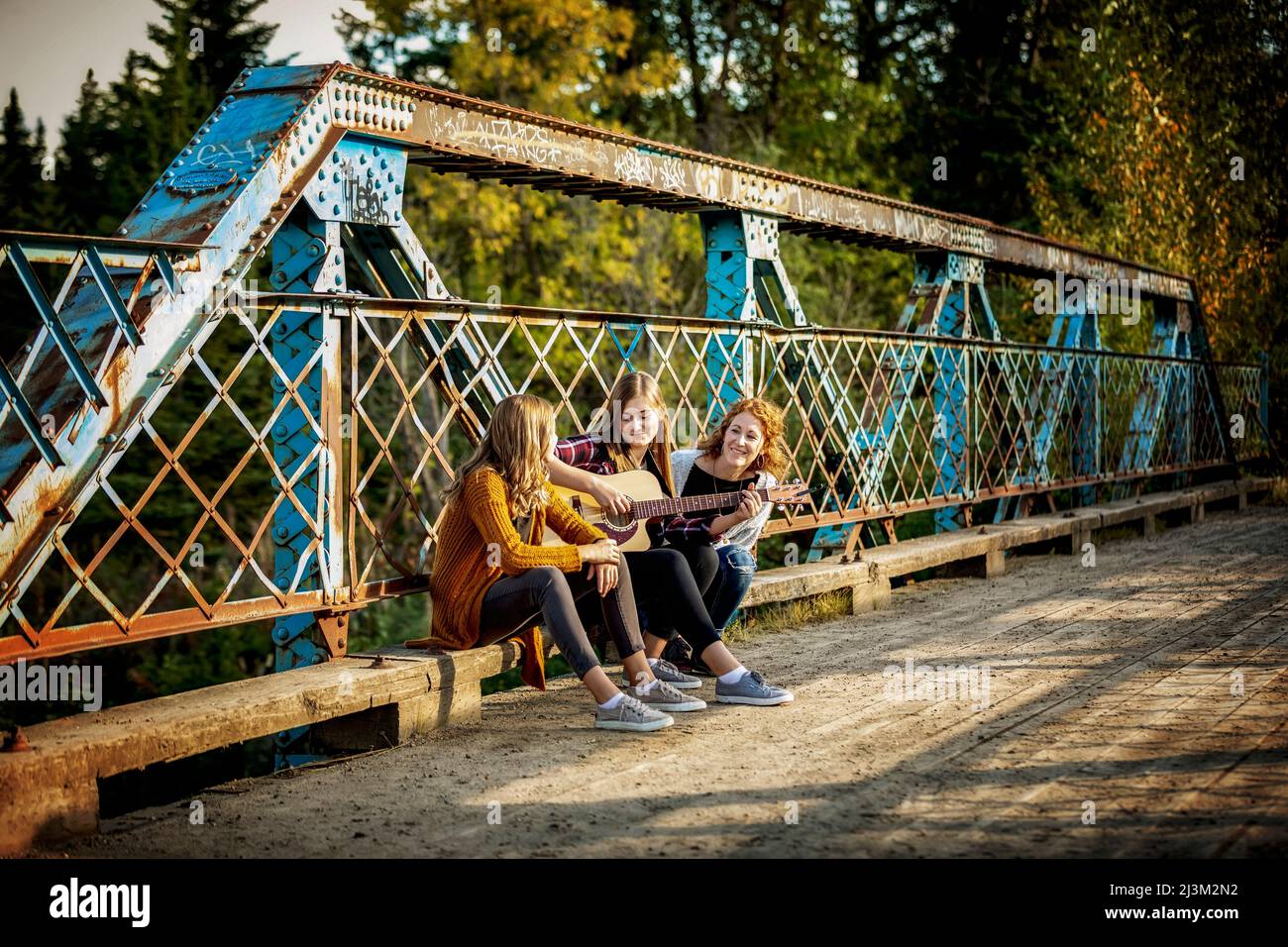 Mutter und zwei Schwestern sitzen auf einer Parkbrücke, eine mit einer akustischen Gitarre, und genießen gemeinsam Musik; Edmonton, Alberta, Kanada Stockfoto