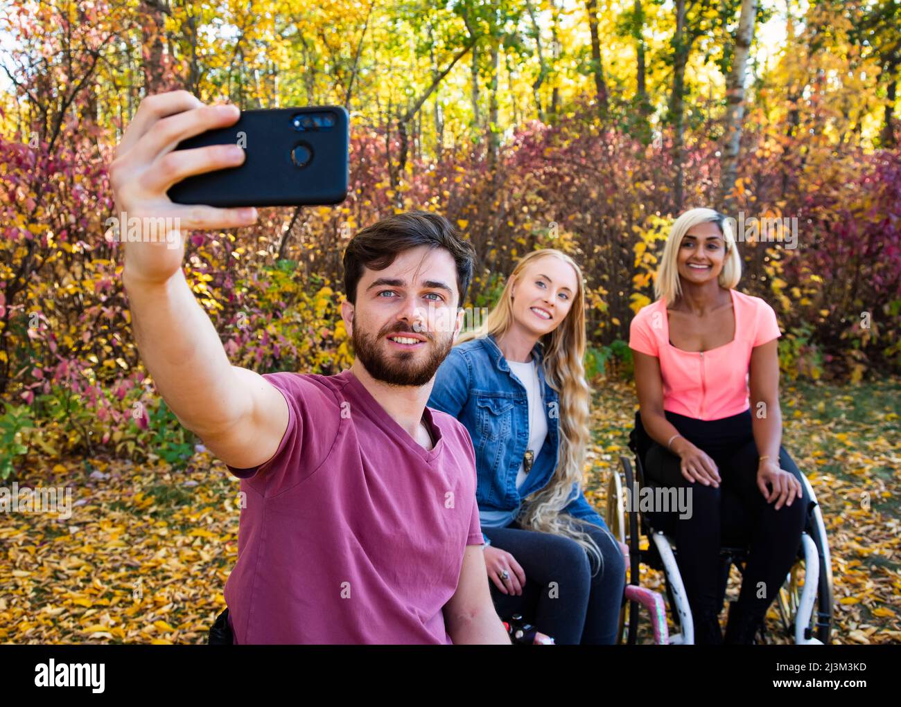 Junge querschnittsgelähmte Männer und Frauen im Rollstuhl, die an einem schönen Herbsttag mit einem Smartphone in einem Park ein Selbstporträt machen Stockfoto