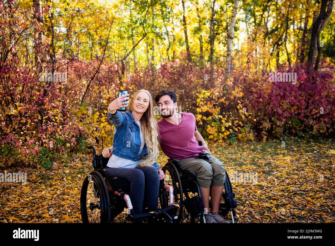 Ein junger querschnittsgelähmter Mann und eine junge Frau im Rollstuhl, die an einem schönen Herbsttag ein Selbstporträt mit einem Smartphone in einem Park machen Stockfoto