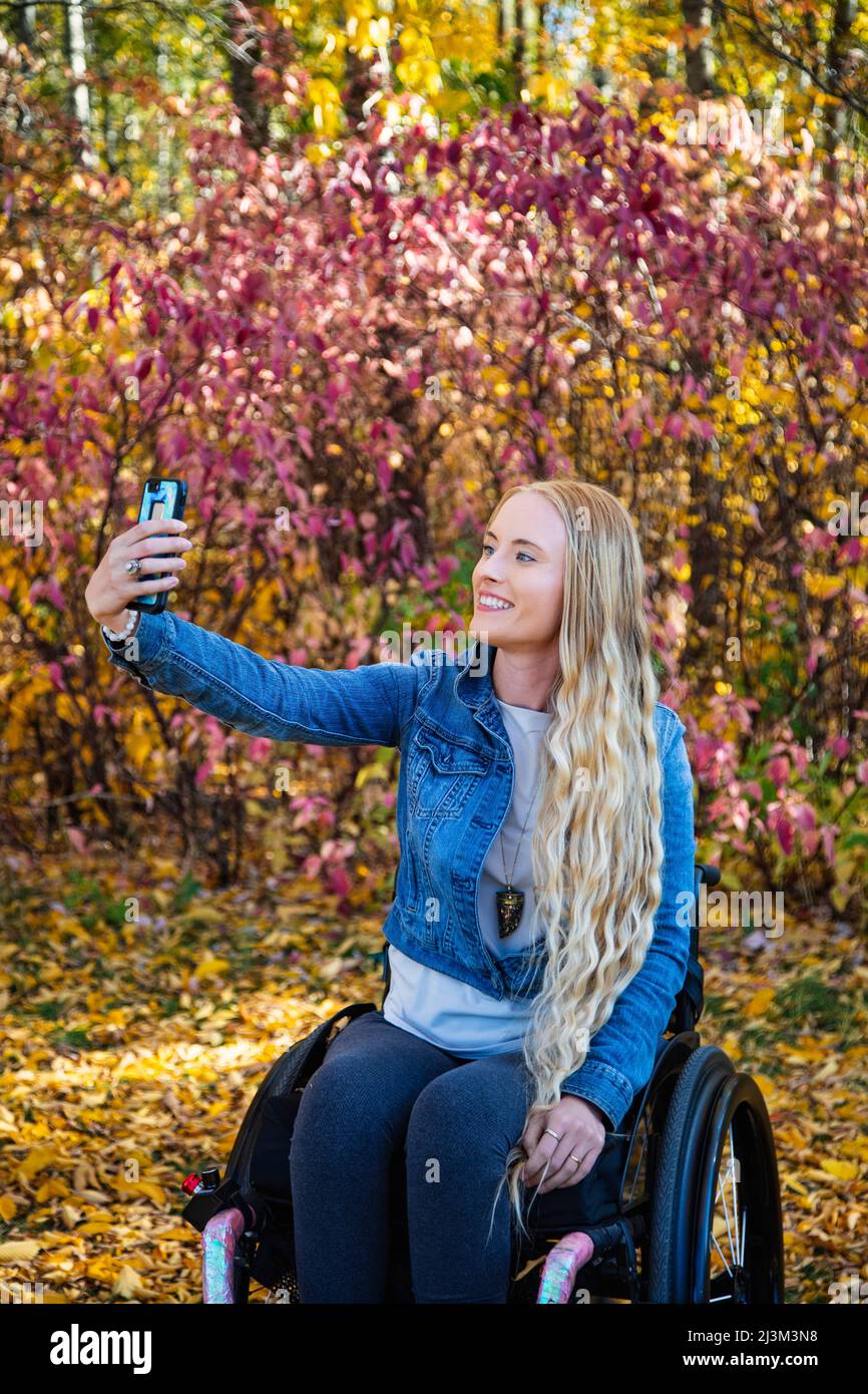 Eine junge querschnittsgelähmte Frau im Rollstuhl, die an einem schönen Herbsttag mit ihrem Smartphone in einem Park ein Selbstporträt macht; Edmonton, Alberta, Kanada Stockfoto