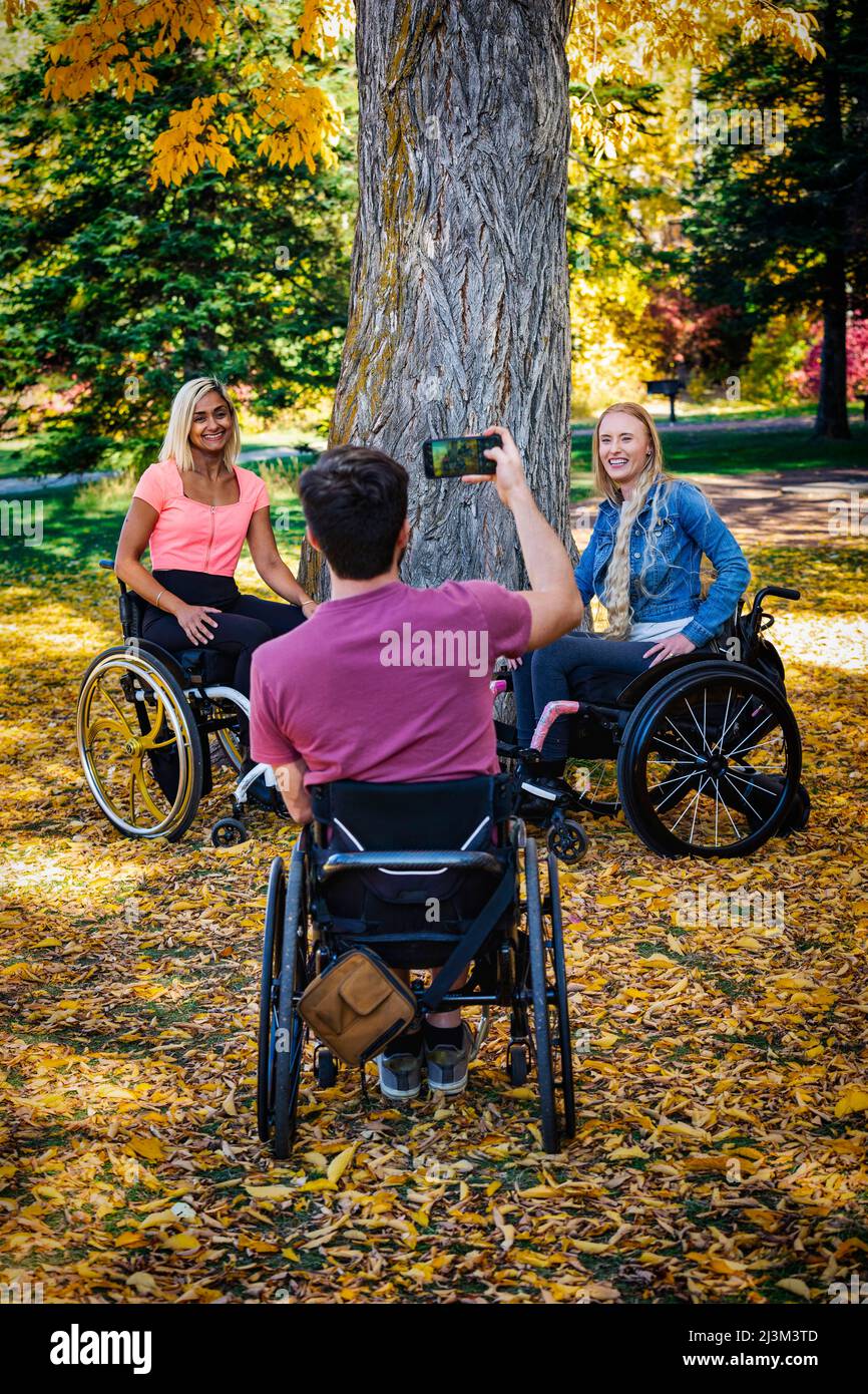 Junge querschnittsgelähmte Männer und Frauen im Rollstuhl, die an einem schönen Herbsttag mit einem Smartphone in einem Park Fotos machen; Edmonton, Alberta, Kanada Stockfoto