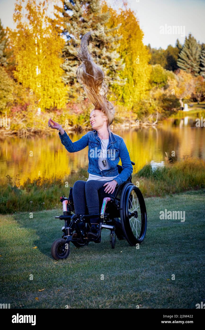 Junge querschnittsgelähmte Frau in ihrem Rollstuhl in einem Park an einem schönen Herbsttag, die sich mit ihrem langen Haar, das nach oben dreht, sorglos fühlt Stockfoto