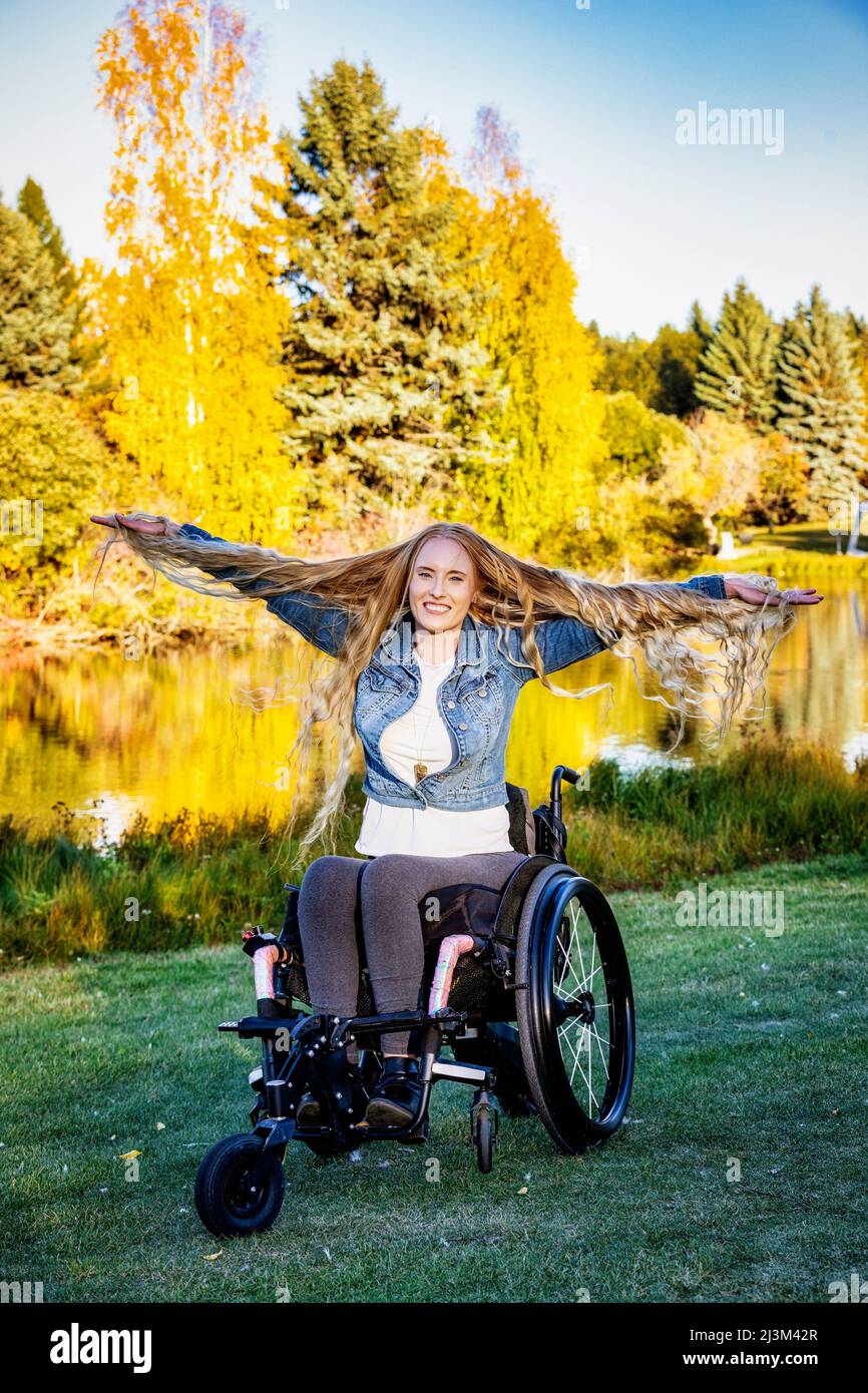 Junge querschnittsgelähmte Frau in ihrem Rollstuhl in einem Park an einem schönen Herbsttag, Gefühl sorglos mit ihren Armen ausgestreckt und zeigt die Länge der h... Stockfoto