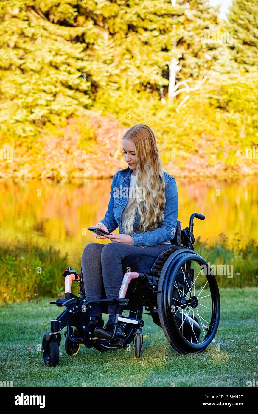 Junge querschnittsgelähmte Frau im Rollstuhl, die an einem schönen Herbsttag in einem Park ein Smartphone benutzt; Edmonton, Alberta, Kanada Stockfoto