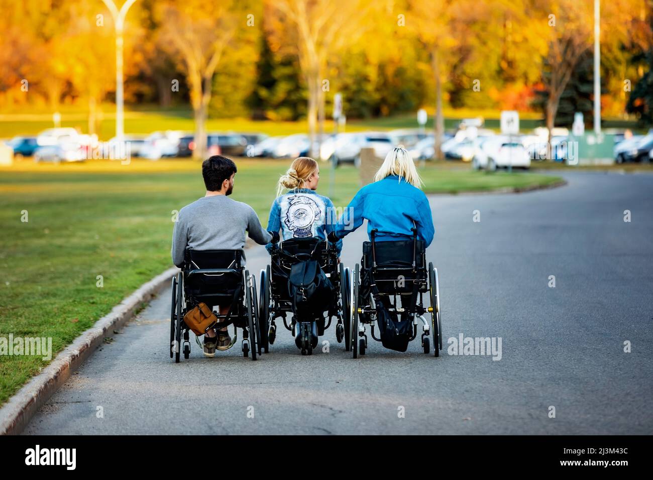 Drei junge Querschnittsgelähmte in ihren Rollstühlen, die an einem schönen Herbsttag durch einen Parkplatz in einem Park gehen und sich in einer Reihe die Hände halten Stockfoto