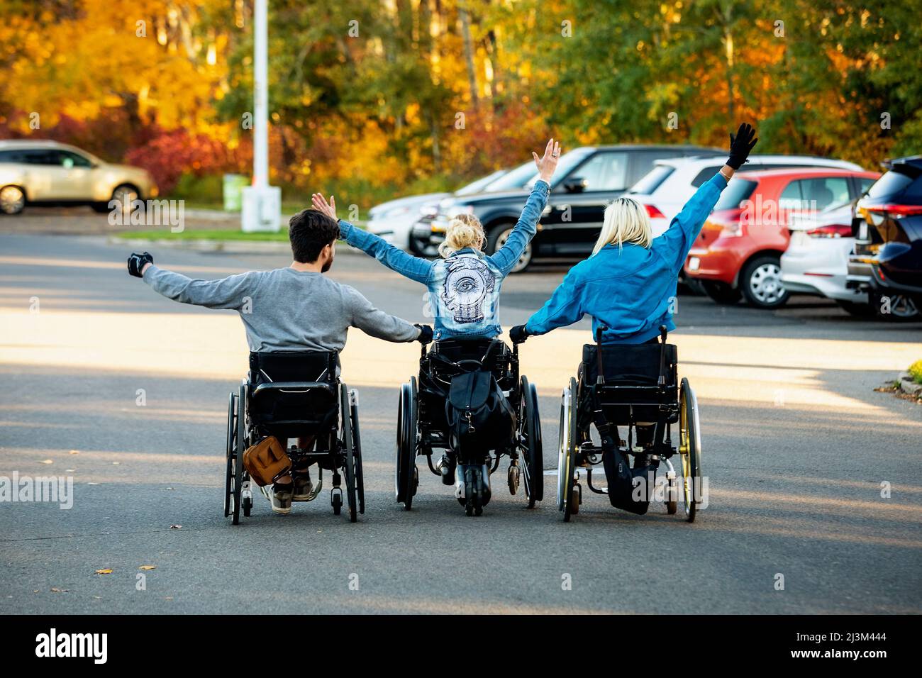 Drei junge querschnittsgelähmte in ihren Rollstühlen, die an einem schönen Herbsttag durch einen Parkplatz in einem Park gehen, mit erhobenen Armen und... Stockfoto