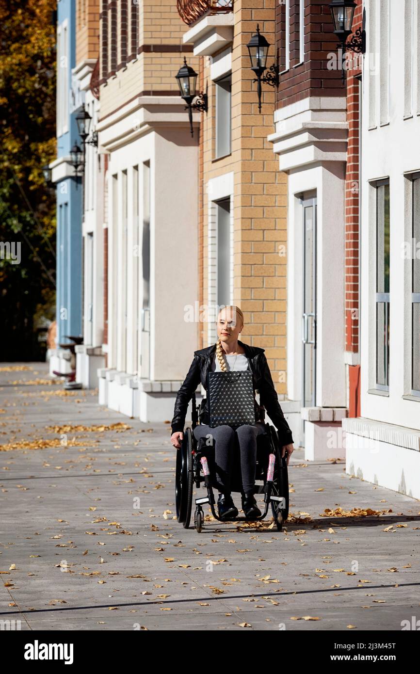 Eine junge querschnittsgelähmte Frau in ihrem Rollstuhl mit einer Einkaufstasche, die an einem schönen Herbsttag einen Stadtgang entlang geht; Edmonton, Alberta, Kanada Stockfoto