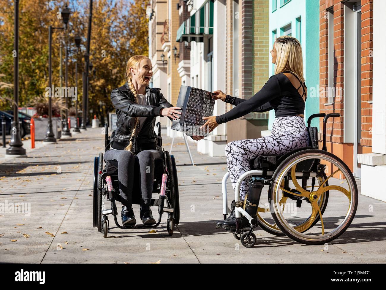 Zwei junge Frauen, die an einem schönen Herbsttag im Rollstuhl paraplegiker sind, besuchen gemeinsam einen Gehweg, einer schenkte dem anderen ein Geschenk Stockfoto