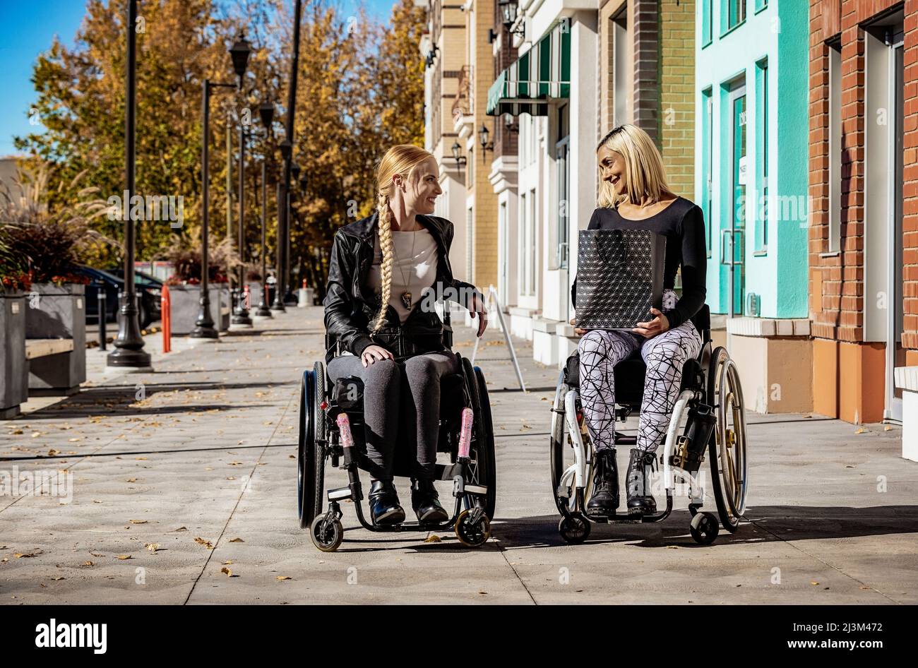 Zwei junge Frauen, die an einem schönen Herbsttag auf einem Gehweg paraplegiker in ihren Rollstühlen besuchten, eine mit einer Einkaufstasche Stockfoto