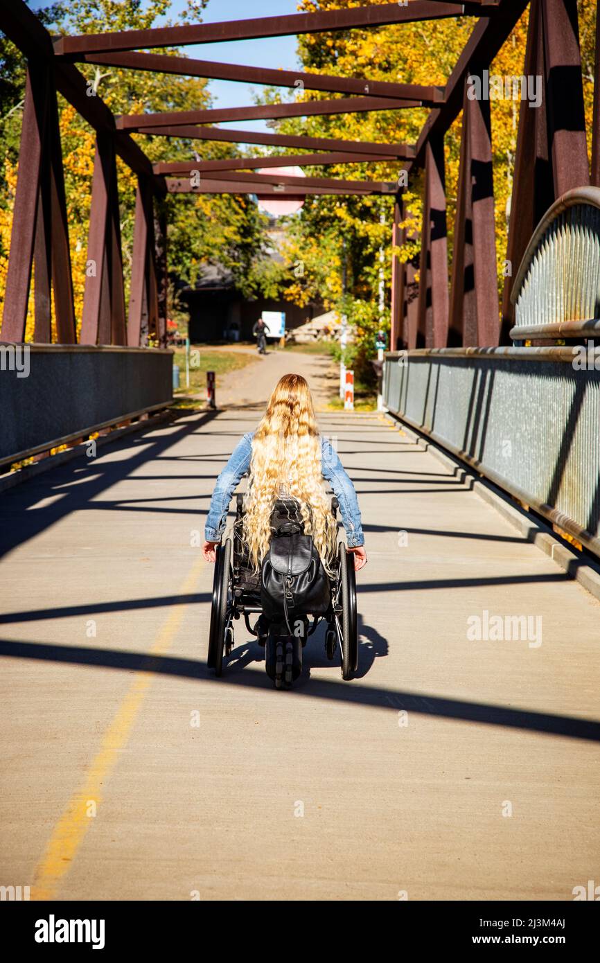 Junge querschnittsgelähmte Frau mit langen blonden Haaren, die an einem schönen Herbsttag mit ihrem Rollstuhl über eine Brücke fährt; Edmonton, Alberta, Kanada Stockfoto