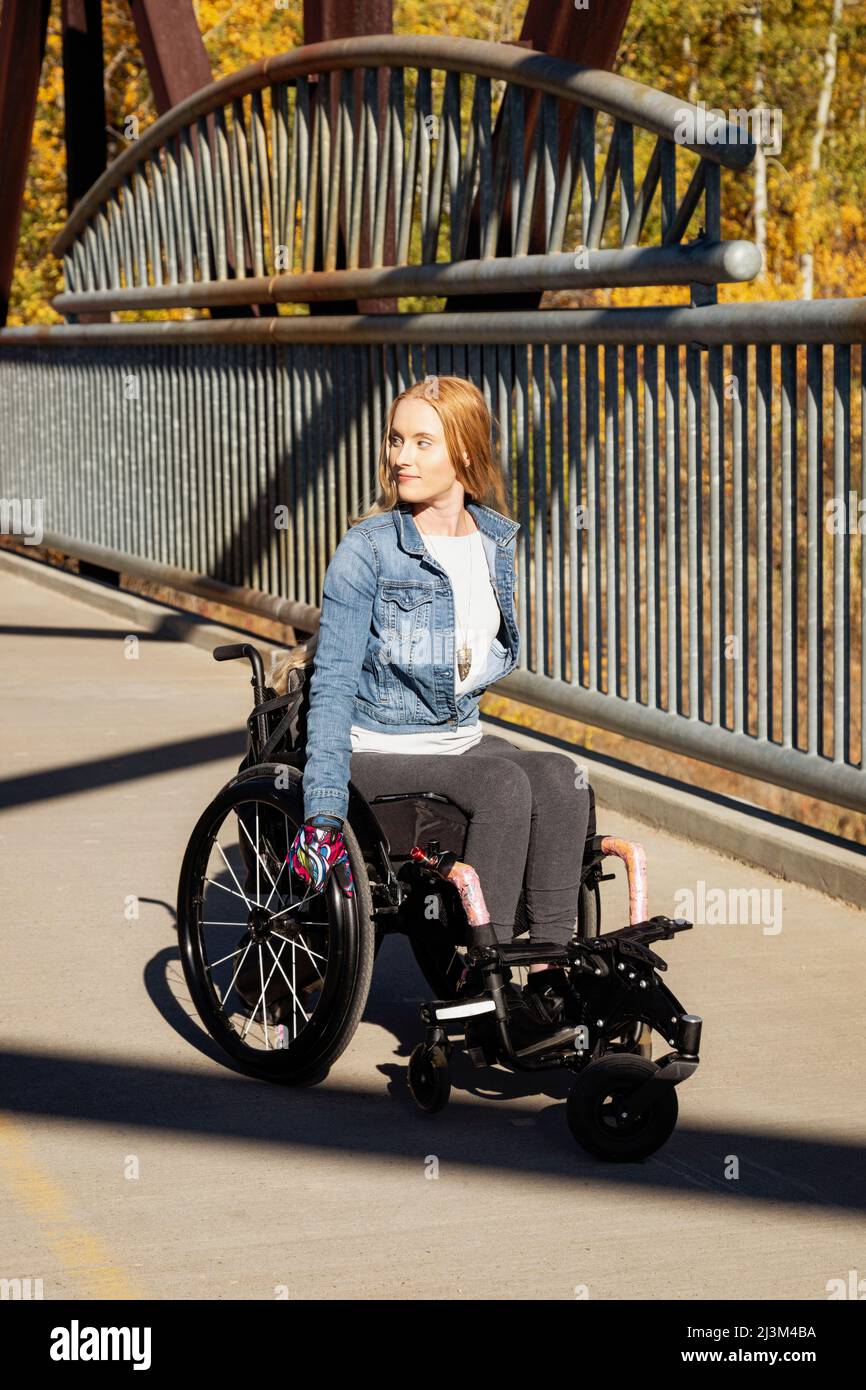Junge querschnittsgelähmte Frau, die an einem schönen Herbsttag mit ihrem Rollstuhl über eine Brücke geht; Edmonton, Alberta, Kanada Stockfoto