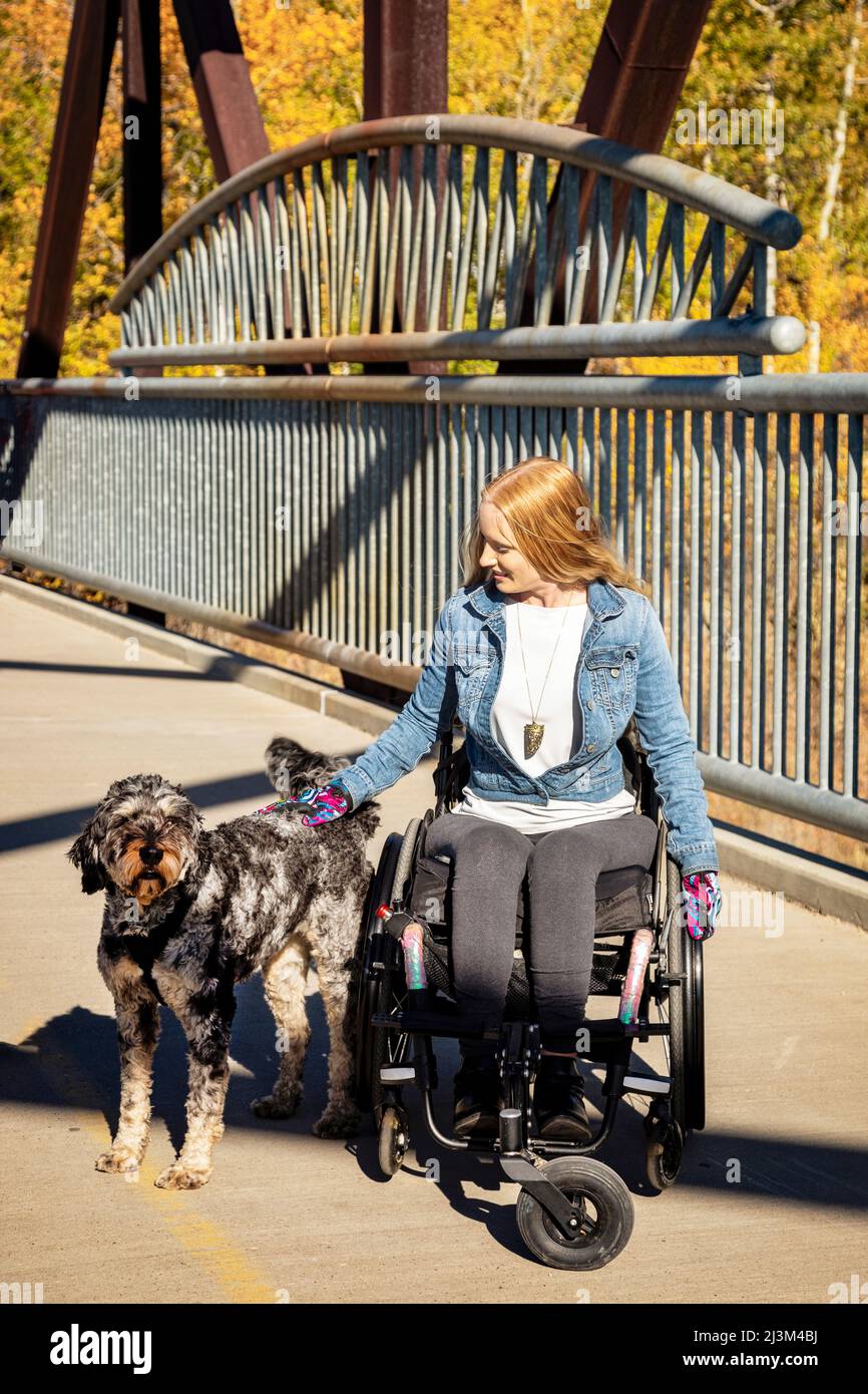 Junge querschnittsgelähmte Frau in ihrem Rollstuhl in einem Park an einem schönen Herbsttag, hält an, um einen Hund auf einer Brücke zu streicheln; Edmonton, Alberta, Kanada Stockfoto