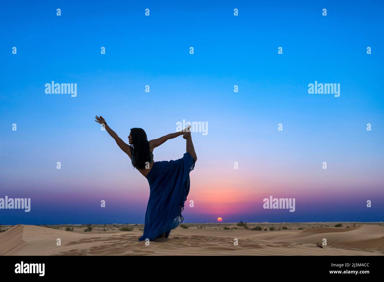 Frau mit langem Rock, hinterleuchtet vom Sonnenuntergang am Wüstensand, stehend in einer Yoga-Tänzerin-Pose; Abu Dhabi, Vereinigte Arabische Emirate Stockfoto