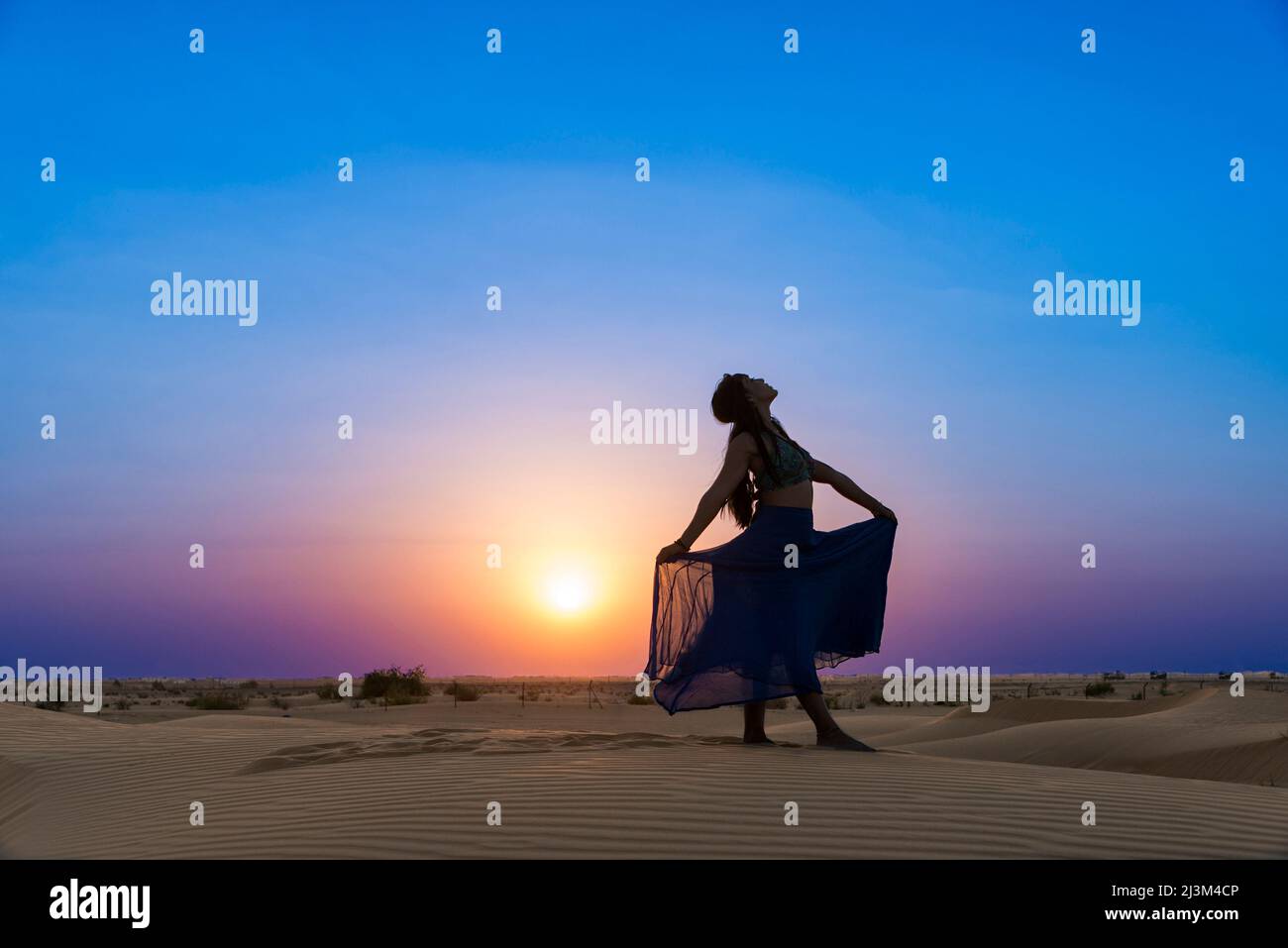 Frau mit langem Rock im Hintergrund der Sonnenuntergang auf dem Wüstensand; Abu Dhabi, Vereinigte Arabische Emirate Stockfoto