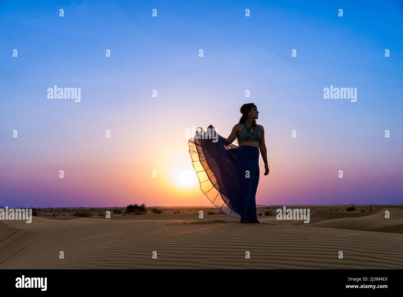 Frau mit langem Rock im Hintergrund der Sonnenuntergang auf dem Wüstensand; Abu Dhabi, Vereinigte Arabische Emirate Stockfoto