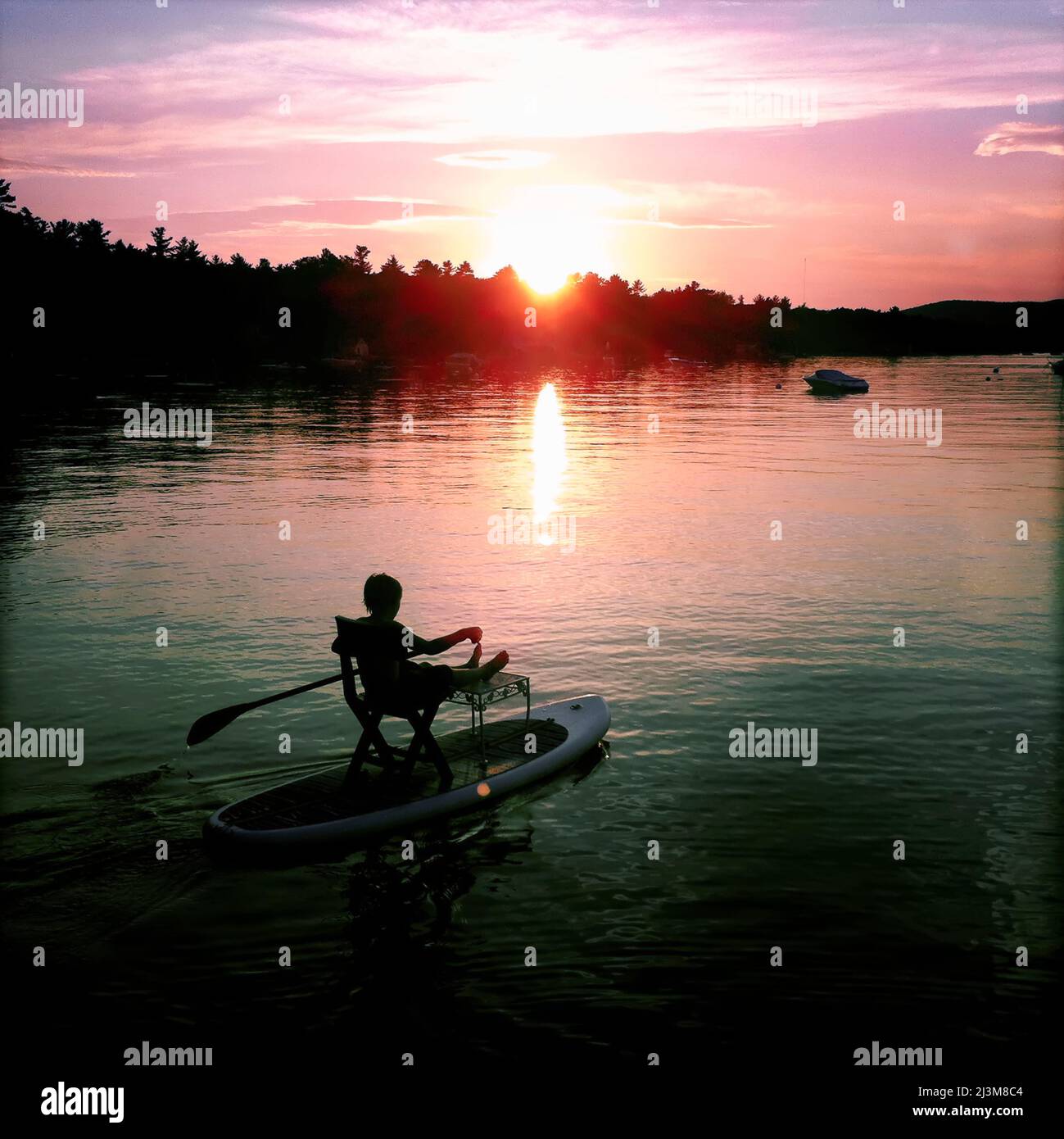 Ein Junge versucht einen setz-down Ansatz auf seinem Stand-up Paddle Board bei Sonnenuntergang.; Sebago Lake, Maine, USA. Stockfoto