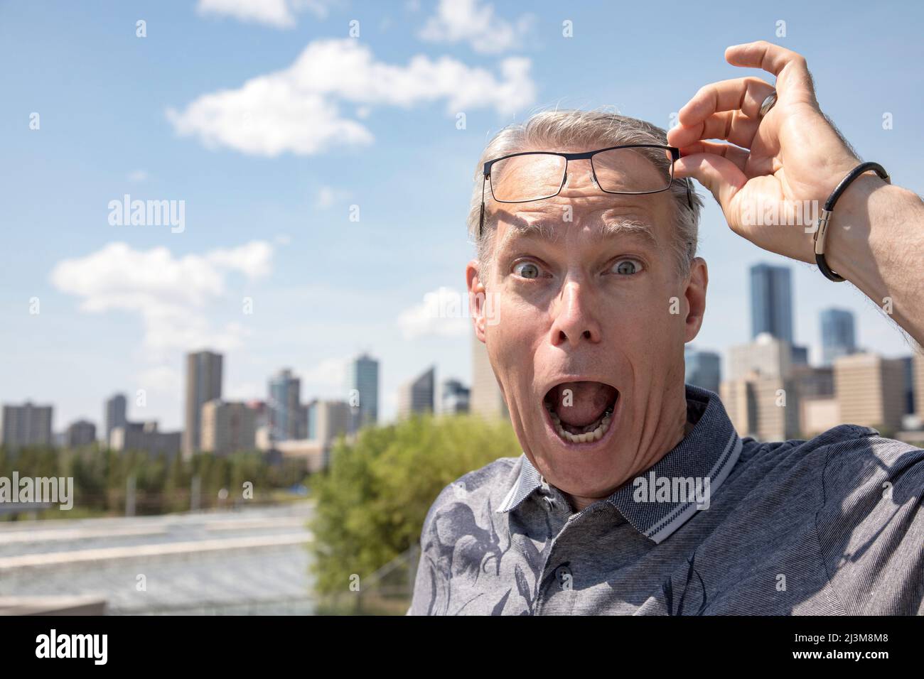 Älterer Mann, der im Freien steht und seine Brille hochhebt, um mit einem überraschenden Gesichtsausdruck auf die Kamera zu blicken; Edmonton, Alberta, Kanada Stockfoto